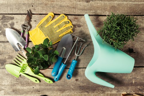 The image shows a selection of gardening tools and supplies arranged on a weathered wooden surface, likely a garden table. Visible items include a small hand trowel, a pair of pruning shears, a yellow gardening glove made of flexible material, a small hand rake, and a set of three miniature gardening tools with blue handles, possibly for planting or soil work. Additionally, there is a light turquoise plastic watering can and a potted plant with dense, dark green foliage, possibly a herb or small shrub, placed on the right side. The background surface shows natural wood grain with some minor weathering, suggesting outdoor use. The scene is well-lit, indicating daytime with natural light, and reflects a tidy and purposeful arrangement of gardening essentials. Such tools and plant care supplies are typical for maintaining a well-kept garden or landscaped outdoor space in Stratford or surrounding areas, supporting the services offered by Landscapers Stratford in garden maintenance, planting, and outdoor improvement projects.