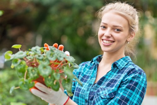 A young woman with blonde hair tied back, wearing a blue plaid shirt and white gardening gloves, is smiling while holding a small potted plant with green leaves and pinkish flowers. She is standing outdoors in a garden environment with a background of blurred greenery consisting of trees and shrubs. The garden appears well-maintained, with a lush appearance typical of a residential outdoor space in Stratford, featuring a variety of planting areas. Natural daylight suggests a clear, mild weather day suitable for outdoor gardening work. The scene reflects a moment of gardening activity, supporting services related to garden planting, landscape design, and outdoor maintenance by Landscapers Stratford, located in Stratford upon Avon and serving local garden renovation needs.