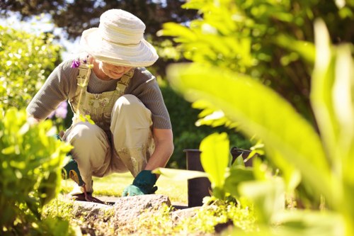 A woman working in a well-maintained front garden with vibrant green foliage, a neatly trimmed hedge in the background, and a grassy lawn area in the foreground. She is wearing a wide-brimmed hat, gardening gloves, and casual outdoor clothing, and is crouched down planting or tending to plants within a flower bed bordered by bricks. The garden features a variety of lush bushes and leafy plants that add colour and texture to the landscape. Potted plants are visible nearby, and the scene is illuminated by natural sunlight, suggesting a bright, clear day, typical of outdoor gardening activities in Stratford. The careful arrangement of plantings and the tidy lawn reflect professional landscaping and garden care services, such as those offered by Landscapers Stratford, focused on creating attractive, sustainable outdoor spaces for homes in the local area.