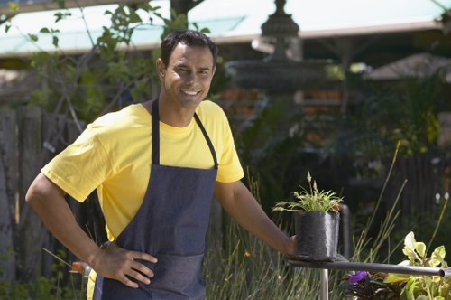 A smiling man wearing a yellow t-shirt and a dark apron stands in a well-maintained garden in Stratford, surrounded by lush green plants and shrubs. In front of him, a simple round table holds a small black plastic pot with young, green foliage. Behind him, there are various garden elements including a wooden fence, some trees, and a decorative garden feature. The garden features a mix of grass, soil, and paving, with a neat border separating the lawn from flower beds and shrub-lined edges. The scene suggests outdoor gardening activities on a bright day, with natural sunlight illuminating the space. The environment reflects typical landscaping and garden maintenance, aligning with services offered by companies like Landscapers Stratford, especially in the context of sustainable gardening practices highlighted on the 'Recycling and Sustainability' page. The setting captures an organized, thriving outdoor space suitable for lawn care, planting, and general garden upkeep in the local area.