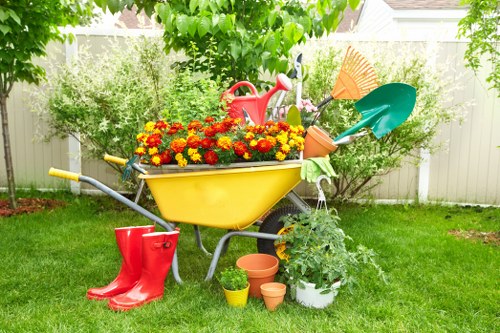 In a lush, well-maintained garden, a bright yellow wheelbarrow is filled with vibrant red and yellow chrysanthemums, positioned on a green lawn with neatly trimmed grass. Surrounding the wheelbarrow are various gardening tools and accessories, including a red watering can placed on top of the flowers, a small green watering can nearby, and several empty terracotta pots of different sizes resting on the ground. A pair of red rubber gardening boots stands adjacent to the wheelbarrow, hinting at recent or upcoming planting activities. Behind, a white garden fence runs horizontally, providing a backdrop for a variety of shrubs and small trees with leafy green foliage, indicating a healthy garden environment. The scene is illuminated by natural daylight, suggesting clear weather, with a few tree branches partially framing the top of the image, contributing to the overall peaceful and productive outdoor atmosphere. This setting reflects professional gardening support, possibly by Landscapers Stratford, in a typical residential garden near Stratford, suitable for lawn and plant care services.