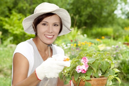 A woman wearing a white hat, gardening gloves, and a light-colored shirt is tending to a potted flowering plant with pink blossoms in an outdoor garden setting. Behind her, there is a well-maintained lawn with dense, green grass, bordered by various shrubs and flowering plants in natural tones of green, pink, and yellow. The garden features a mix of flower beds and paved pathways, with some trees and hedges providing a lush backdrop. The scene appears to be in daylight, under mild outdoor conditions, with soft natural light illuminating the garden and the woman's activity. This image exemplifies typical gardening work within a landscaped outdoor space, reflecting services offered by licensed landscapers in Stratford or similar locations, with attention to plant care and outdoor maintenance.