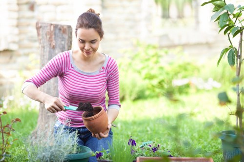 A woman with dark hair tied back, wearing a pink and white striped long-sleeve shirt, is tending to plants in a well-maintained garden on a bright day. She is holding a small terracotta pot filled with soil in one hand and using a gardening tool in the other to work with the soil. In front of her, there are several small potted plants with purple and green foliage arranged on a low garden table. The garden area features a lush green lawn, a tree trunk visible in the background, and a variety of flowering plants and shrubs along a fence or stone boundary wall. The scene is illuminated by natural daylight, suggesting pleasant weather, ideal for outdoor gardening activities, which could be part of a gardening service in Stratford or surrounding areas provided by Landscapers Stratford.