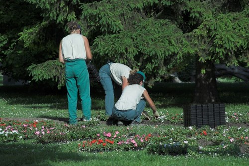 The image depicts three individuals working in a well-maintained garden area, located in Stratford, with a large, dense green tree providing shade in the background. Two of the people are crouched down near flower beds that feature a variety of small, colorful flowers, such as pink, red, and white blooms, arranged along the edge of a lush, neatly trimmed grass lawn. The third person stands nearby, wearing a sleeveless top and teal trousers, observing or assisting with the gardening activities. The garden surface includes a combination of grass, flowering borders, and possibly some soil or mulch around the plants. The overall outdoor setting appears tidy and cared for, with the activity taking place under natural daylight, suggesting a clear weather condition suitable for gardening services. This scene highlights typical landscape maintenance or planting tasks associated with professional gardening work in Stratford, aligning with services offered by Landscapers Stratford in lawn and flower bed care.