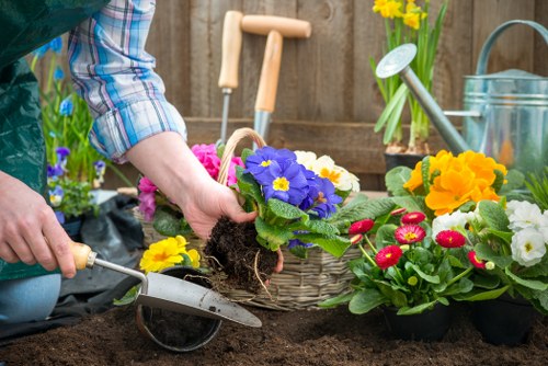 A person wearing a plaid shirt is gardening in a backyard garden, preparing soil for planting colorful flowers such as primroses and other bedding plants. The gardener's hands are visible as they place a potted flowering plant into the dark, well-tilled soil, which appears rich and moist. In the background, a wooden fence surrounds the space, with garden tools including a small trowel and watering can resting nearby on a garden table or surface. Bright yellow daffodils grow in a pot on the table, adding vibrant colour to the scene, while other potted plants ready for planting are positioned around the workspace. The garden features a mix of lush greenery, flowering plants, soil beds, and garden accessories, creating a well-maintained outdoor space typical of Stratford gardens. The scene is set on a slightly overcast day, with natural light illuminating the area, highlighting the natural tones of earth, leaves, and blossoms, reflecting professional gardening and landscaping practices that Landscapers Stratford could undertake to enhance gardens across Stratford and the surrounding postcode areas.