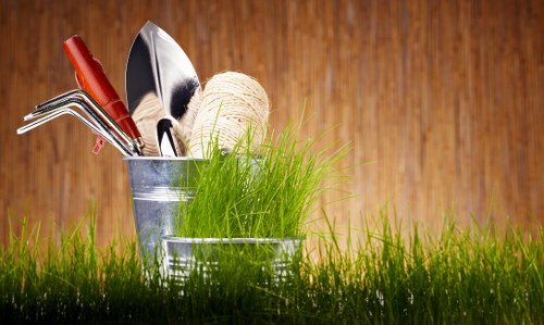 The image shows a close-up of a lush, green lawn with well-maintained grass in the foreground. Behind the grass, there are two small, silver metal containers holding gardening tools and supplies. One container contains a pair of pruning shears with red handles, a small trowel, and a gardening fork, while the other holds a spool of twine and a bunch of fresh green grass or seedlings. The background features a warm-toned wooden surface, possibly a garden shed or fence, providing a natural backdrop. The overall scene highlights outdoor gardening preparation and equipment, typical of residential or community garden settings in Stratford and surrounding areas, aligning with professional gardening practices offered by Landscapers Stratford. The lighting suggests a bright, possibly sunny day, emphasizing the vibrant colours and textures of the tools and greenery. This setup is indicative of organized garden maintenance activities and the tools used in planting and landscaping services within the Stratford area.