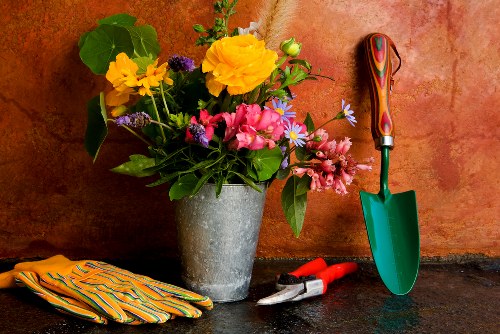 A close-up image of a garden workspace featuring a grey concrete flower pot filled with a colourful arrangement of fresh flowers, including yellow, pink, and purple blossoms, set against a textured terracotta wall. Surrounding the pot are gardening tools and accessories: a pair of red-handled pruning shears, a small trowel with a green handle, a pair of patterned gardening gloves, and an orange and yellow striped cloth. The scene is illuminated by natural light, highlighting the vibrant colours of the flowers and tools, and suggesting an outdoor or well-lit garden maintenance area in Stratford. The arrangement reflects typical gardening activities related to planting, tending, and caring for garden plants, with a focus on floral displays and tools for outdoor garden upkeep, aligning with the services offered by Landscapers Stratford in the local area.