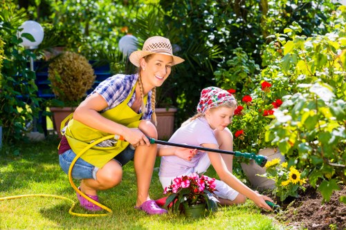 In a lush backyard garden, a woman and a young girl are engaged in gardening activities amidst vibrant plants and flowers. The woman, dressed in a checkered shirt, denim shorts, pink gardening gloves, and a straw hat, is kneeling on the grass while holding a yellow garden hose. The girl, wearing a rain hat with a floral pattern, a pink T-shirt, and pink shoes, is seated on the grass and tending to a flower bed with pink and yellow blooms, using her hand to gently plant or water the soil. The garden setting features a neatly maintained lawn with bright green grass, edged by a variety of flowering plants, including roses and sunflowers, close to a paved stone pathway. In the background, there are dense trees and shrubs creating a natural enclosure, with some garden lights visible hanging from the foliage, suggesting a well-cared-for outdoor space typical of Stratford gardens. The scene captures a sunny day with natural light illuminating the vibrant colours and textures of the garden, reflecting a peaceful, family-friendly outdoor environment where gardening services could enhance outdoor beauty and functionality.