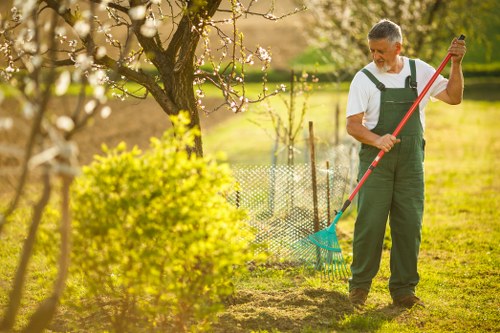 A middle-aged man wearing green gardening overalls, a white T-shirt, and black gloves is raking soil in a lush, well-maintained backyard garden on a bright, sunny day. The garden features a neatly mown lawn with dense, green grass in the foreground, bordered by a flower bed with small, yellow and pink flowering plants. Tall, blooming trees with pink blossoms dominate the background, casting dappled sunlight across the area. A low, wire fence runs along the edge of the garden, separating it from adjacent landscaped spaces. The scene captures the outdoor environment in spring or early summer, with vibrant natural tones of green, yellow, and pink, highlighting a typical residential garden managed by local landscapers in Stratford. The image underscores early-stage garden maintenance, possibly part of ongoing lawn and shrubcare services offered by Landscapers Stratford, with a focus on outdoor beautification and safety practices in outdoor work environments.